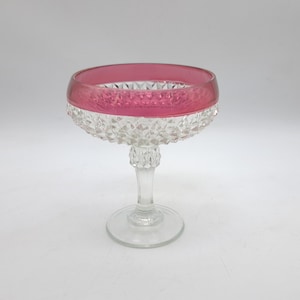 Peut inclure: Une coupe sur pied en verre transparent avec un large bord rose foncé et un motif en losanges texturé sur l'extérieur du bol et sur le pied. L'objet repose sur une base circulaire, présentant une combinaison de surfaces en verre lisse et facetté.
