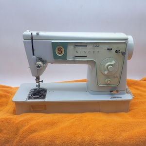 Puede incluir: Una máquina de coser Singer vintage en acabado blanco roto con un panel de control verde pálido. La máquina presenta un dial circular de longitud de puntada, varias palancas de ajuste deslizantes y el logotipo de Singer en rojo y dorado.