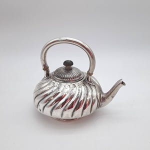 May include: A silver teapot with a curved handle and a spout. The body of the teapot has a ridged, swirling design. The lid is topped with a small, dark knob. The teapot is set against a white background.