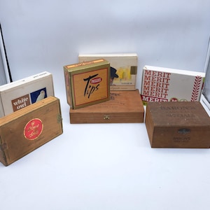 May include: An assortment of vintage boxes, including cigar boxes and cigarette packs. The boxes are made of wood and cardboard, with various printed labels and text, such as "White Owl", "Phillies Tips", and "Merit".