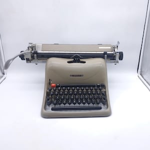 May include: A vintage beige typewriter with black keys and a silver carriage. The typewriter has a red button and a black handle. The brand name "LEADER" is visible on the front. The machine is set against a white background.