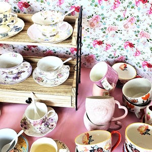 May include: A collection of teacups and mugs in various floral patterns and colors, displayed on a two-tiered wooden stand and a pink surface. Some of the cups have handles, while others have heart-shaped handles. The cups are arranged in a decorative and eye-catching way.