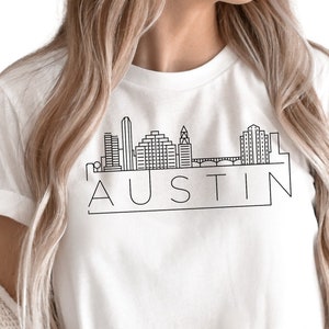 K&ouml;nnte beinhalten: Wei&szlig;es T-Shirt mit einer schwarzen Umrandung der Skyline von Austin, Texas und dem Wort "AUSTIN" in einem schwarzen Kasten.