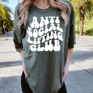 K&ouml;nnte beinhalten: Ein gr&uuml;nes T-Shirt mit dem Text "Anti Social Lifting Club" in wei&szlig;er Schrift im Retro-Stil.