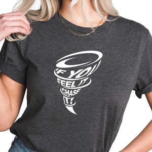May include: A dark gray t-shirt with a white graphic of a tornado with the text "If you feel it chase it!" inside.