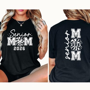 Op de afbeelding: Zwart T-shirt met witte tekst die "Senior Mom 2026" op de voorkant en "Senior Mom 2026" met een cheerleading pompom-afbeelding op de achterkant zegt.