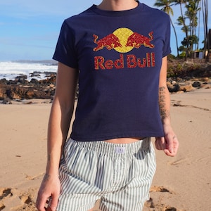 A person wearing a navy blue t-shirt with a Red Bull logo and a graphic of two red bulls on a yellow background, standing on a beach with palm trees in the background.