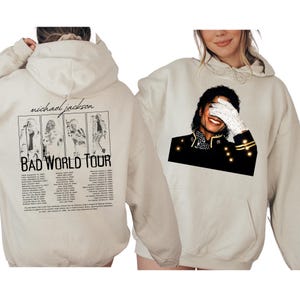May include: Beige hoodie featuring a graphic of Michael Jackson. The front displays a portrait of Jackson, while the back has tour images and text that reads "BAD WORLD TOUR". The hoodie has a front pocket and a drawstring hood.