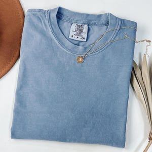 May include: A folded, dusty blue Comfort Colors t-shirt with a gold necklace featuring a pendant. The shirt's tag is visible, and a brown hat is partially seen in the upper left corner. Dried leaves are in the lower right corner.