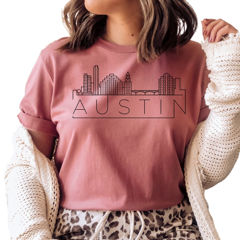 K&ouml;nnte beinhalten: Ein pinkfarbenes T-Shirt mit einer schwarzen Umrandung der Skyline von Austin, Texas und dem Wort "AUSTIN" darunter.
