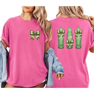 May include: A pink t-shirt featuring a green bow on the front and three green Sprite bottles with bows on the back. The bottles have yellow labels. The shirt has short sleeves and a relaxed fit.