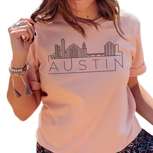 K&ouml;nnte beinhalten: Ein pinkfarbenes T-Shirt mit einer schwarzen Umrandung der Skyline von Austin, Texas und dem Wort "AUSTIN" in einem Kasten darunter.