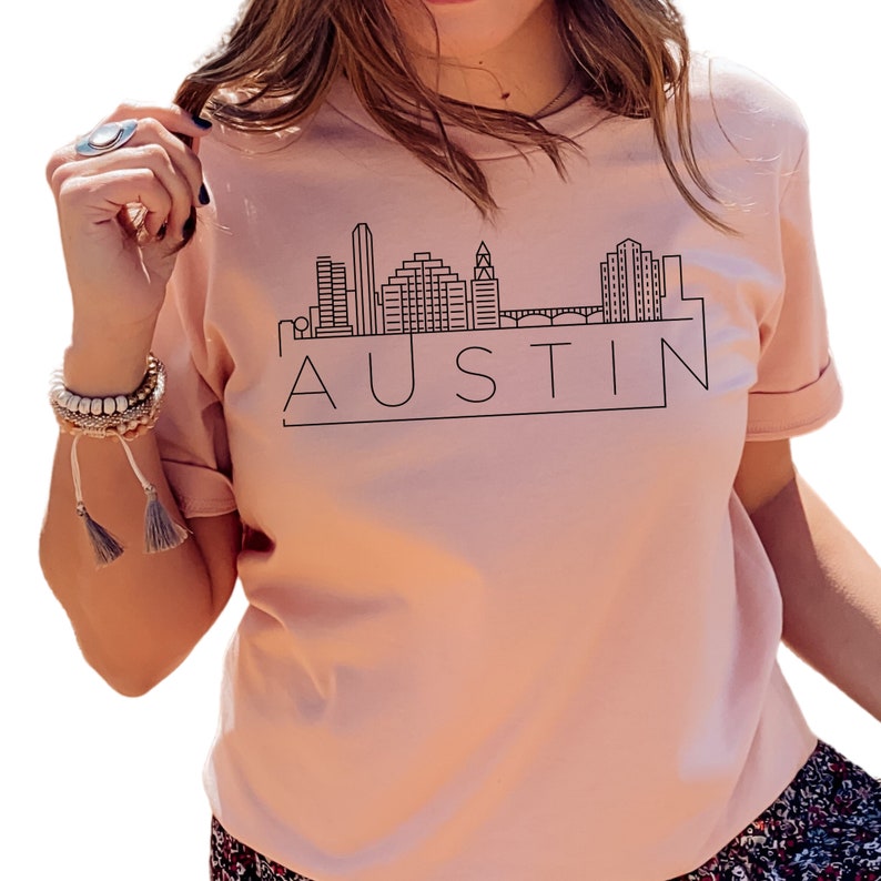 K&ouml;nnte beinhalten: Ein pinkfarbenes T-Shirt mit einer schwarzen Umrandung der Skyline von Austin, Texas und dem Wort "AUSTIN" in einem Kasten darunter.