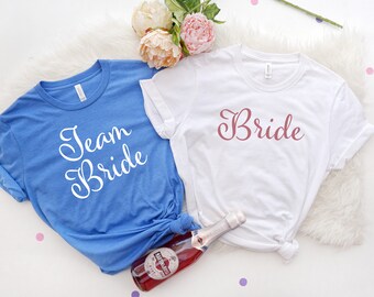 May include: Two white t-shirts with text on them. One shirt says "Team Bride" in white text on a blue shirt. The other shirt says "Bride" in pink text on a white shirt.