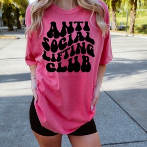 K&ouml;nnte beinhalten: Ein pinkfarbenes T-Shirt mit einer schwarzen Grafik, auf der in einer Retro-Schrift "Anti Social Lifting Club" steht.
