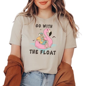 May include: Beige t-shirt with the text "GO WITH THE FLOAT". The graphic features a skeleton relaxing on a pink flamingo floatie, holding a drink. The design includes blue water, birds, and a yellow shirt.