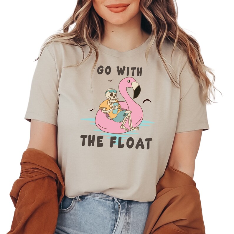May include: Beige t-shirt with the text "GO WITH THE FLOAT". The graphic features a skeleton relaxing on a pink flamingo floatie, holding a drink. The design includes blue water, birds, and a yellow shirt.