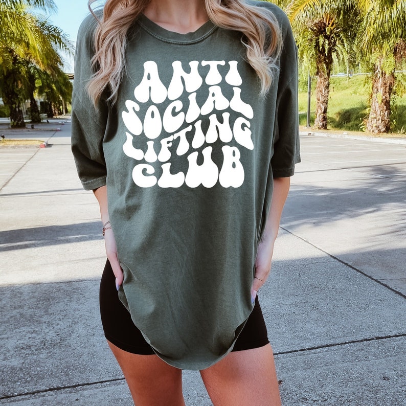K&ouml;nnte beinhalten: Ein gr&uuml;nes T-Shirt mit dem Text "Anti Social Lifting Club" in wei&szlig;er Schrift im Retro-Stil.