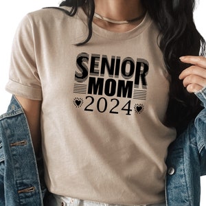 May include: A beige t-shirt with black text that reads "Senior Mom 2024" with a heart on either side of the year.