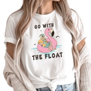May include: White t-shirt with a graphic of a skeleton relaxing on a pink flamingo float. The skeleton is wearing sunglasses and holding a drink. The text "GO WITH THE FLOAT" is above and below the graphic.