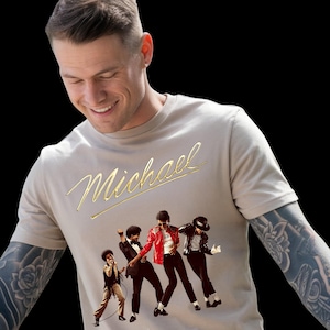 May include: A beige t-shirt featuring the name "Michael" in gold script above a graphic of Michael Jackson in various iconic poses. The graphic includes images of Michael Jackson in a red jacket, a black jacket, and other outfits.