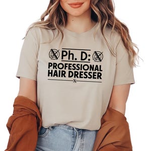 May include: A beige t-shirt with a black graphic that reads "Ph. D.: Professional Hair Dresser" with a pair of scissors and a comb graphic on either side of the text.