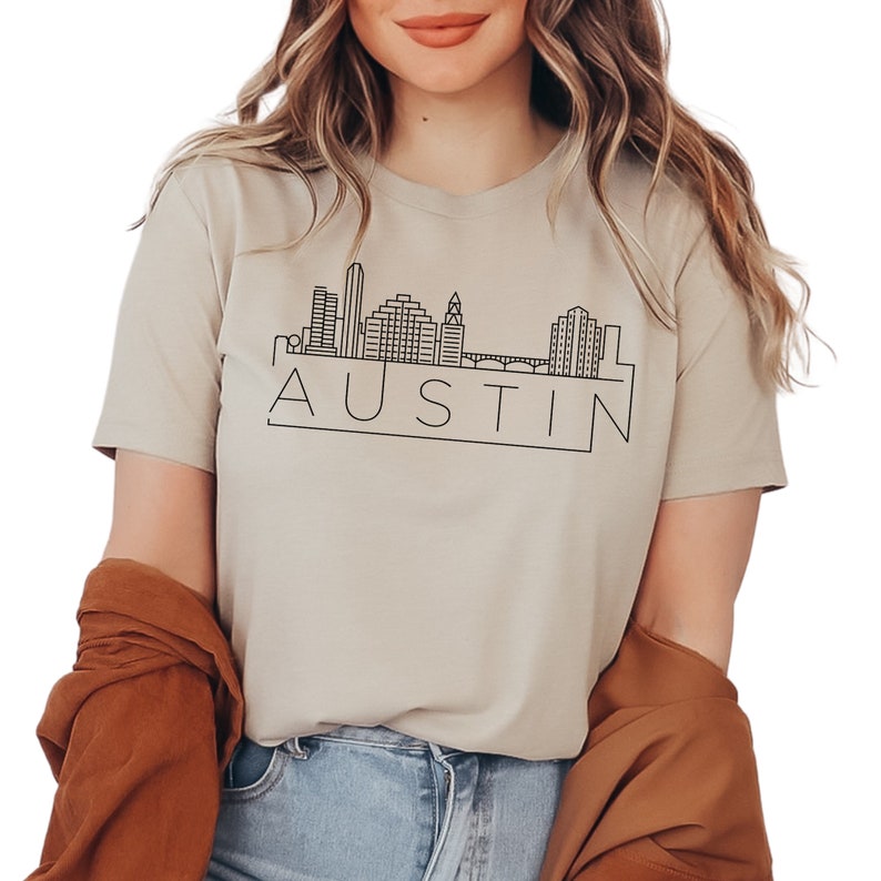 K&ouml;nnte beinhalten: Ein beiges T-Shirt mit einer schwarzen Umrandung der Skyline von Austin, Texas und dem Wort "AUSTIN" darunter.