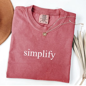 May include: A dusty rose Comfort Colors t-shirt with the word "simplify" printed in white. A gold-colored necklace with a pendant rests on the shirt. A brown hat and dried plant are in the background.