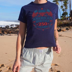 A person wearing a navy blue t-shirt with an American Heritage 250 years design standing on a beach.