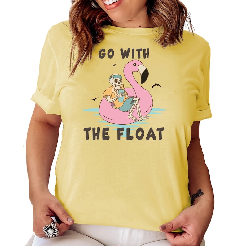 May include: Yellow t-shirt with the text "GO WITH THE FLOAT". The graphic shows a skeleton relaxing on a pink flamingo floatie, holding a drink. The design includes birds and water, creating a fun, summery vibe.