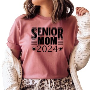 May include: A pink t-shirt with black text that reads "Senior Mom 2024" with a heart on either side of the year.