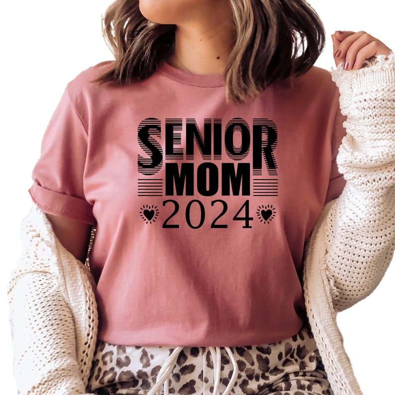 May include: A pink t-shirt with black text that reads "Senior Mom 2024" with a heart on either side of the year.