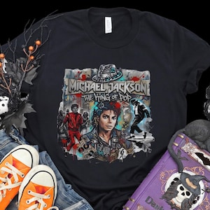 May include: Black t-shirt featuring a graphic of Michael Jackson with the text "Michael Jackson The King of Pop." The design includes images of Jackson in various poses, a fedora, and a colorful background.