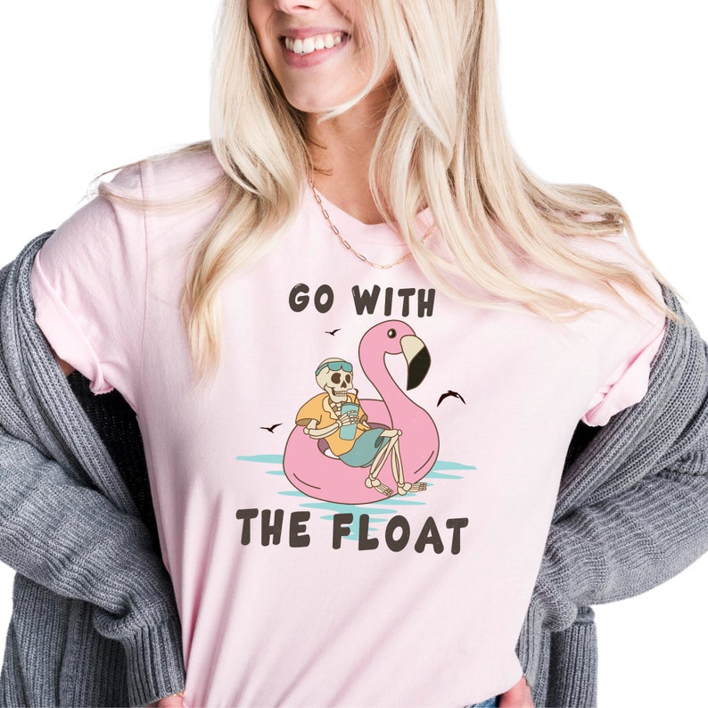 May include: A light pink t-shirt featuring a skeleton relaxing on a pink flamingo float in the water. The skeleton is wearing sunglasses and holding a drink. The text "GO WITH THE FLOAT" is printed on the shirt.