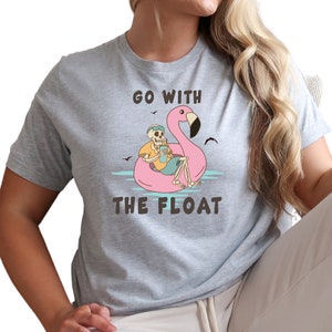 May include: A light gray t-shirt featuring a skeleton relaxing on a pink flamingo float in the water. The skeleton wears sunglasses and holds a drink. The text "GO WITH THE FLOAT" is printed on the shirt.