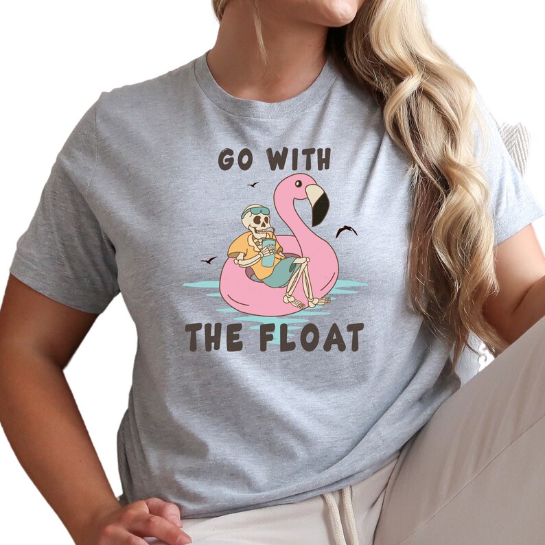 May include: A light gray t-shirt featuring a skeleton relaxing on a pink flamingo float in the water. The skeleton wears sunglasses and holds a drink. The text "GO WITH THE FLOAT" is printed on the shirt.