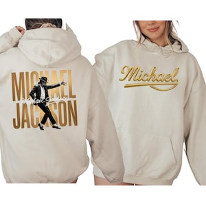 May include: Cream-colored hoodie featuring a gold-colored graphic of Michael Jackson on the back, with his name and image. The front of the hoodie has the name "Michael" in gold script. The hoodie has a front pocket and a hood.