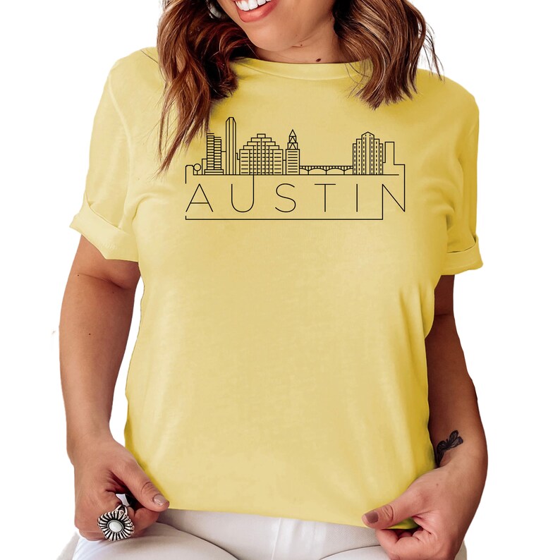 K&ouml;nnte beinhalten: Ein gelbes T-Shirt mit einer schwarzen Umrandung der Skyline von Austin, Texas und dem Wort "AUSTIN" auf der Vorderseite.