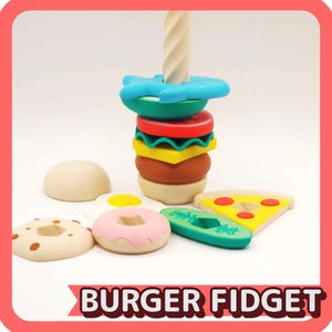 May include: A colorful burger-themed fidget toy with a variety of food-shaped pieces, including a burger, pizza slice, donut, and avocado. The burger has a wooden dowel through the center. The text "BURGER FIDGET" is at the bottom.