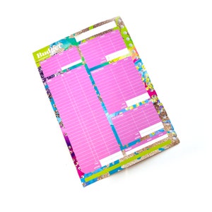 May include: A pink budget planner with a colourful floral design and gold glitter accents. The planner has sections for income, expenses, savings, and debt.