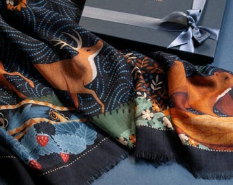 May include: A black and blue patterned scarf with a deer and a fox design. The scarf is folded and displayed on a blue surface with a black gift box with the text "Le Chale Bleu France" on the top.