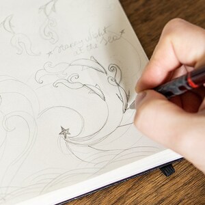 Puede incluir: Un dibujo a l&aacute;piz de delfines, olas y una estrella en un cuaderno de papel blanco. El texto "Starry night at the Sea" est&aacute; escrito a mano encima del dibujo. El cuaderno est&aacute; abierto sobre una superficie de madera.