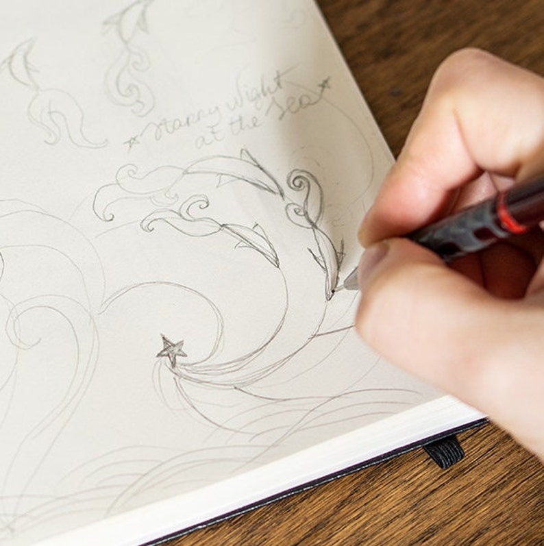 Puede incluir: Un dibujo a l&aacute;piz de delfines, olas y una estrella en un cuaderno de papel blanco. El texto "Starry night at the Sea" est&aacute; escrito a mano encima del dibujo. El cuaderno est&aacute; abierto sobre una superficie de madera.