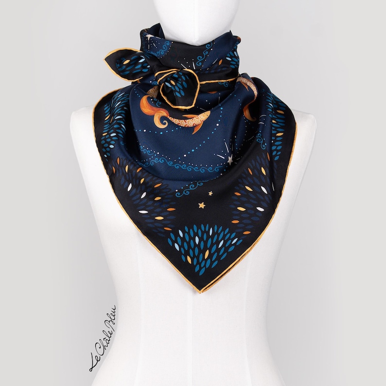 Puede incluir: Un pa&ntilde;uelo cuadrado de seda con fondo azul marino y borde dorado. El pa&ntilde;uelo presenta un dise&ntilde;o caprichoso con una sirena, estrellas y follaje estilizado en tonos naranja, blanco y azul. El pa&ntilde;uelo se exhibe en un maniqu&iacute;.
