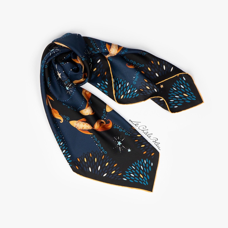 Puede incluir: Pa&ntilde;uelo de seda azul marino con un dise&ntilde;o celestial. El pa&ntilde;uelo presenta detalles dorados, blancos y turquesas, con un borde y el texto "Le Ch&acirc;le Bleu" en blanco. El pa&ntilde;uelo est&aacute; doblado y drapeado.