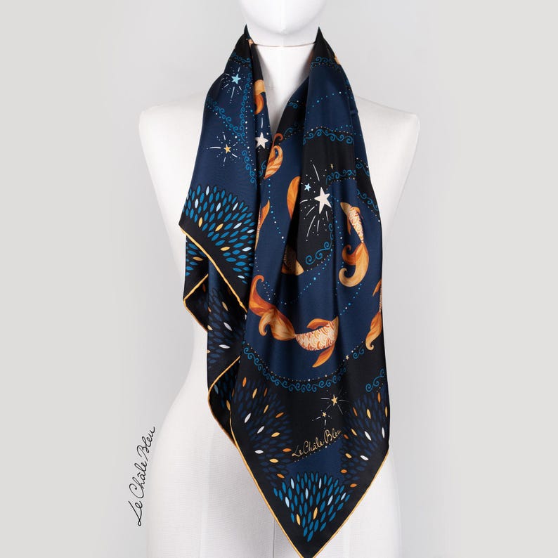 Puede incluir: Un pa&ntilde;uelo de seda azul marino con un dise&ntilde;o celestial, con peces dorados, estrellas y elementos florales estilizados. El pa&ntilde;uelo tiene un borde negro con ribete dorado y el texto "Le Chale Bleu".