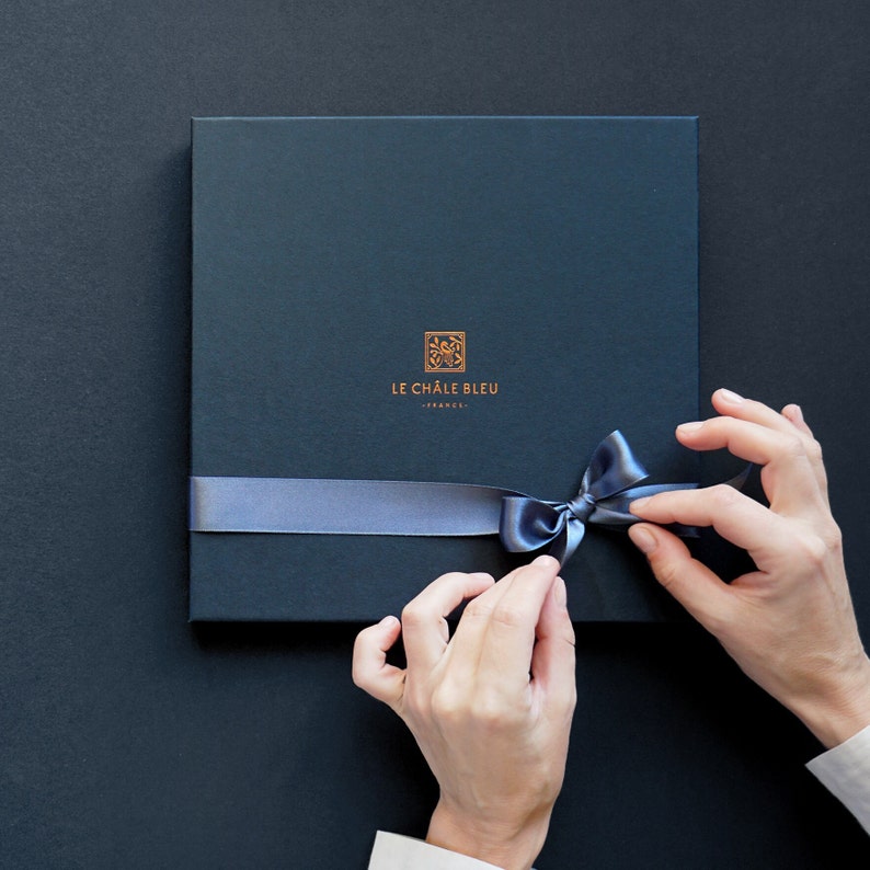 Puede incluir: Una caja de regalo azul marino con una cinta de raso y un lazo a juego. La caja tiene el texto dorado "LE CH&Acirc;LE BLEU - FRANCE". Se ven manos atando el lazo.