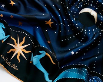 May include: A navy blue silk scarf with a celestial design. The scarf features illustrations of stars, a sun, and blue deer-like creatures. The text "Le Chale Bleu" is printed on the scarf. The scarf has a gold border.