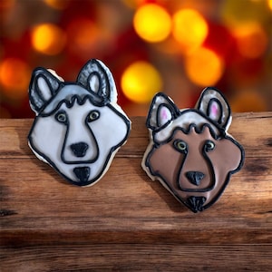 Puede incluir: Dos galletas con forma de lobo con glaseado blanco, gris y marrón, delineadas en negro. Las galletas están sobre una superficie de madera con un fondo borroso de luces amarillas y rojas. Las galletas tienen rasgos faciales detallados.