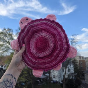 May include: A handmade crochet turtle in shades of pink and magenta. The turtle has a round shell with concentric circles of color, four pink feet, and a smiling face. The background is a blue sky with clouds.